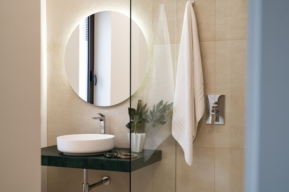 Un baño moderno con un espejo redondo, un lavamanos blanco, una encimera verde, una planta en maceta y una toalla colgada en la pared.