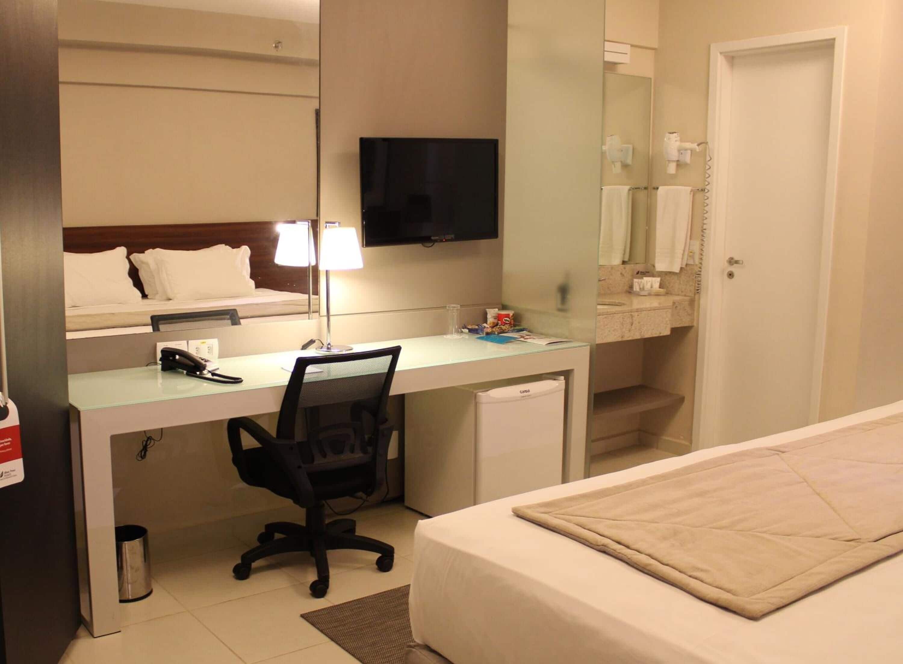 A hotel room with a bed, a desk with a chair, a television, a mirror, and a bathroom with a towel rack.