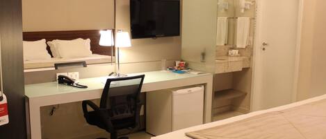 A hotel room with a bed, a desk with a chair, a television, a mirror, and a bathroom with a towel rack.