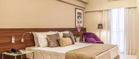 A hotel room with a large bed, a bedside table with a phone, a chair, and a window with curtains.