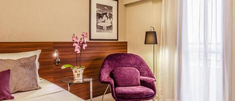 A hotel room with a bed, a purple armchair, a small table with a vase of flowers, a framed picture on the wall, and a window with sheer curtains.