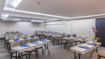 A conference room with rows of tables and chairs, air conditioning units on the ceiling, and a door on the right.