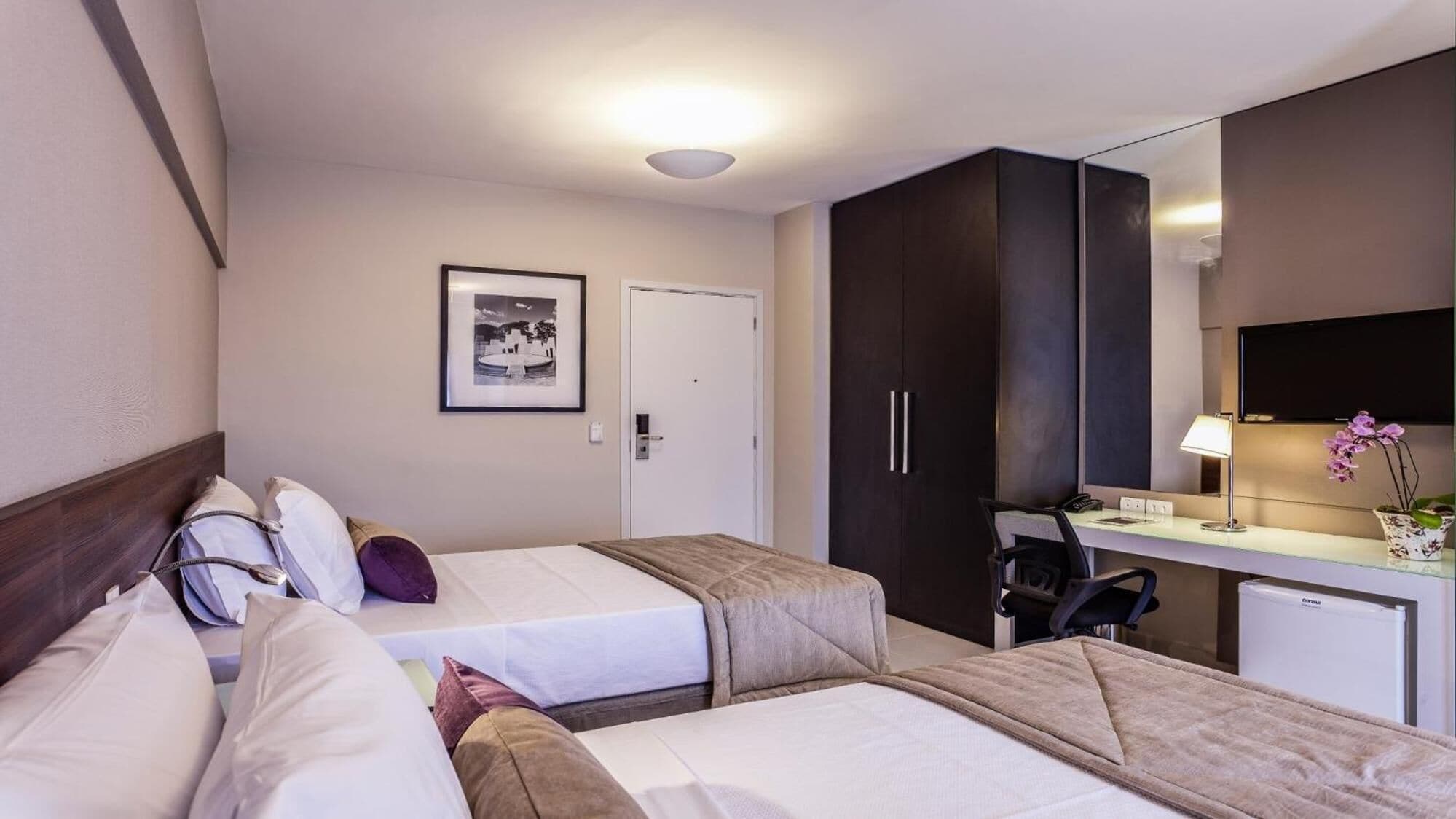 A hotel room with two beds, a desk with a chair, a mirror, a framed picture on the wall, and a door.
