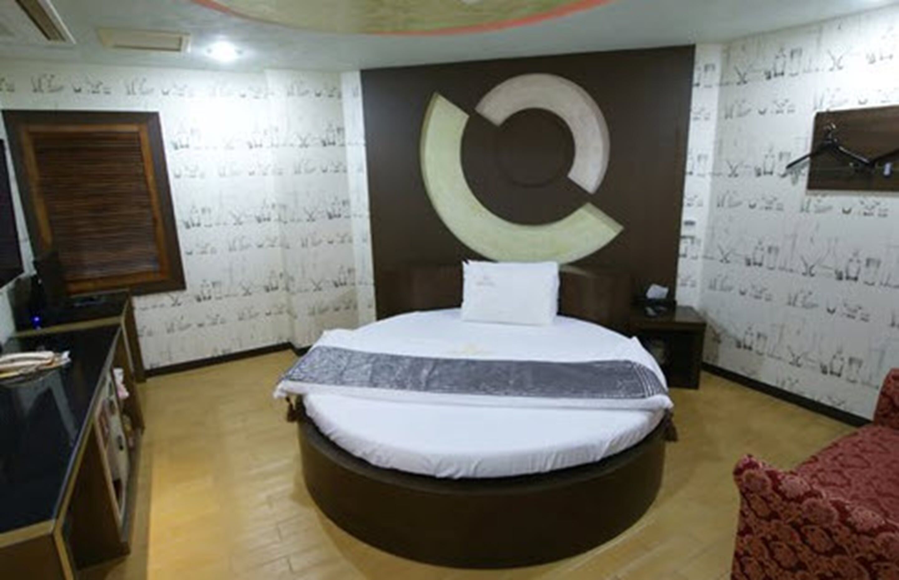 Una cama redonda en una habitación con papel tapiz estampado, suelo de madera y una silla roja.
