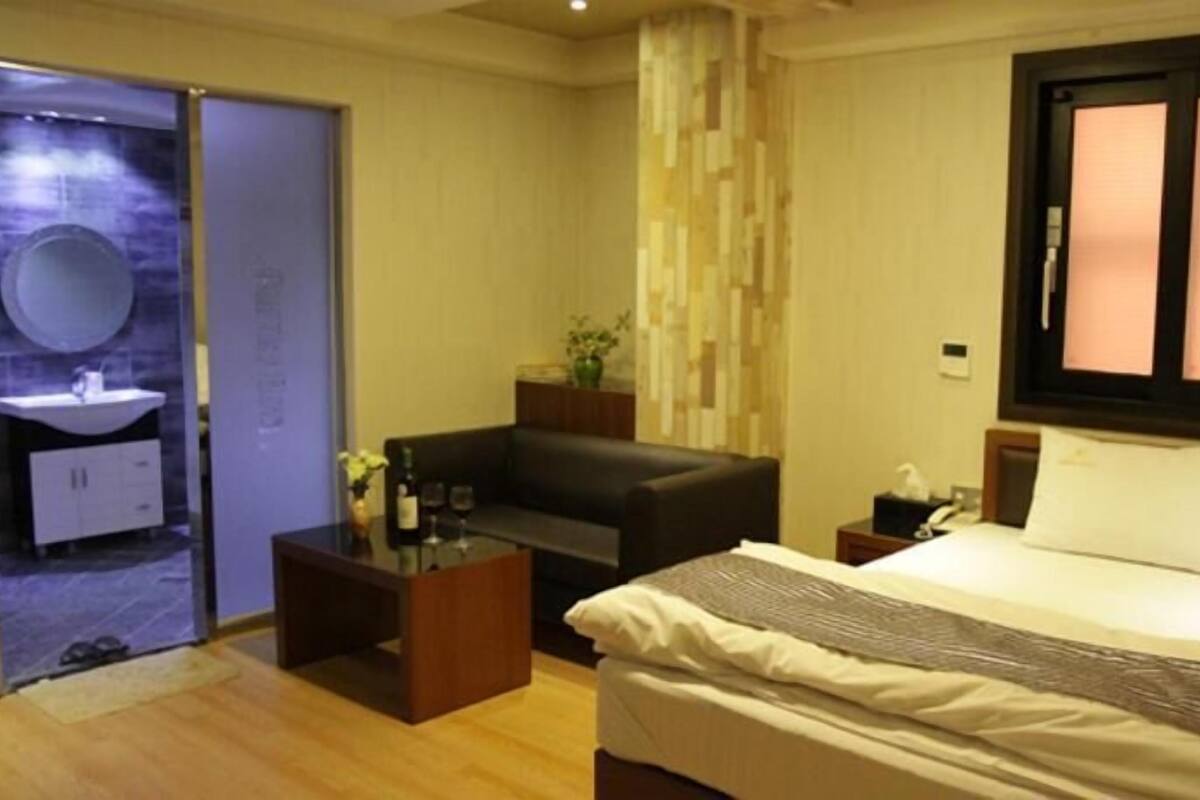 A hotel room with a bed, a sofa, a small table, a mirror, and a window with blinds.