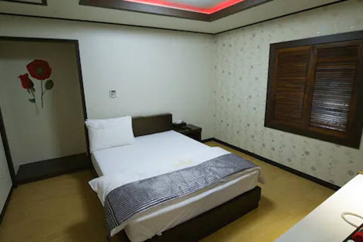A bedroom with a bed, a nightstand, a wall clock, a window with blinds, and a door with a rose decoration.