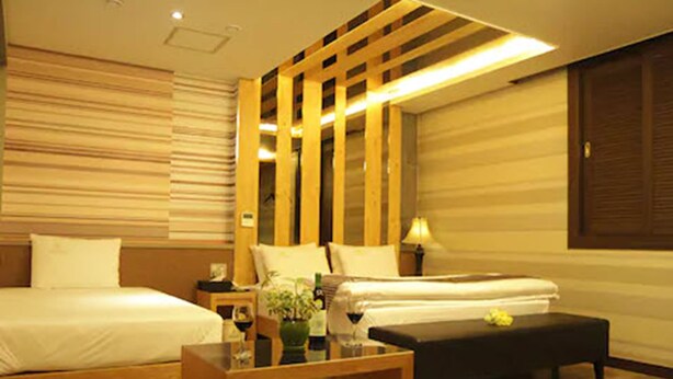 A hotel room with two beds, a wooden bench, a small table with a vase and wine glasses, and a wall-mounted lamp. - Goryeo Hotel (Andong)