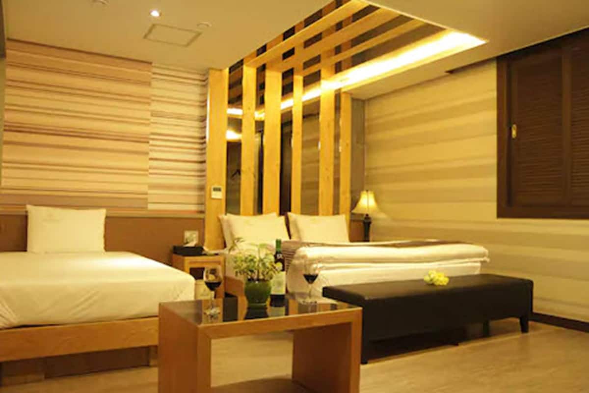 A hotel room with two beds, a wooden bench, a small table with a vase and wine glasses, and a wall-mounted lamp.