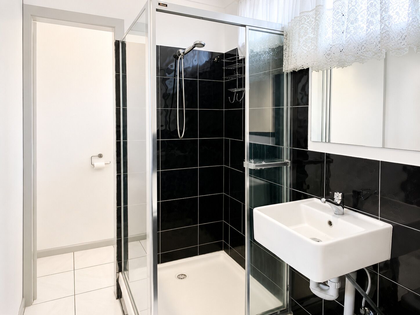 Un baño moderno con cabina de ducha de vidrio, un lavabo blanco y paredes cubiertas de azulejos negros.