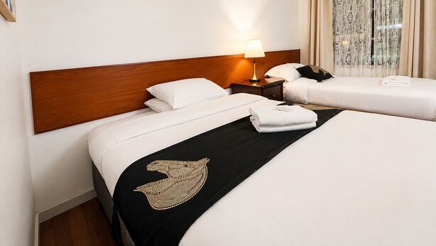A hotel room with two beds, a wooden headboard, a nightstand with a lamp, and a window with curtains. - Beaconsfield Lodge Motel (Beaconsfield)