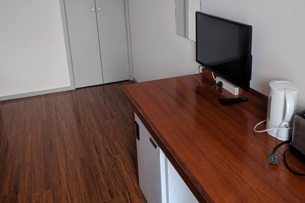 A room with a wooden desk, a television, a coffee maker, and a white appliance, likely a mini-fridge or microwave.