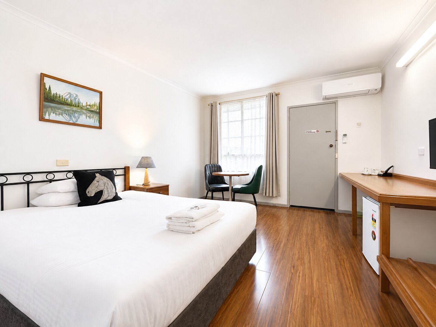 A hotel room with a bed, a desk, a chair, a window with curtains, an air conditioner, and a painting on the wall.