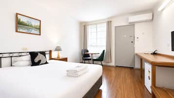 A hotel room with a bed, a desk, a chair, a window with curtains, an air conditioner, and a painting on the wall.