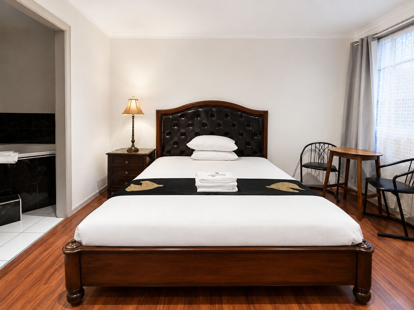 A neatly made bed with a dark headboard, a bedside lamp, a small table with chairs, and a view of the outside through a window.