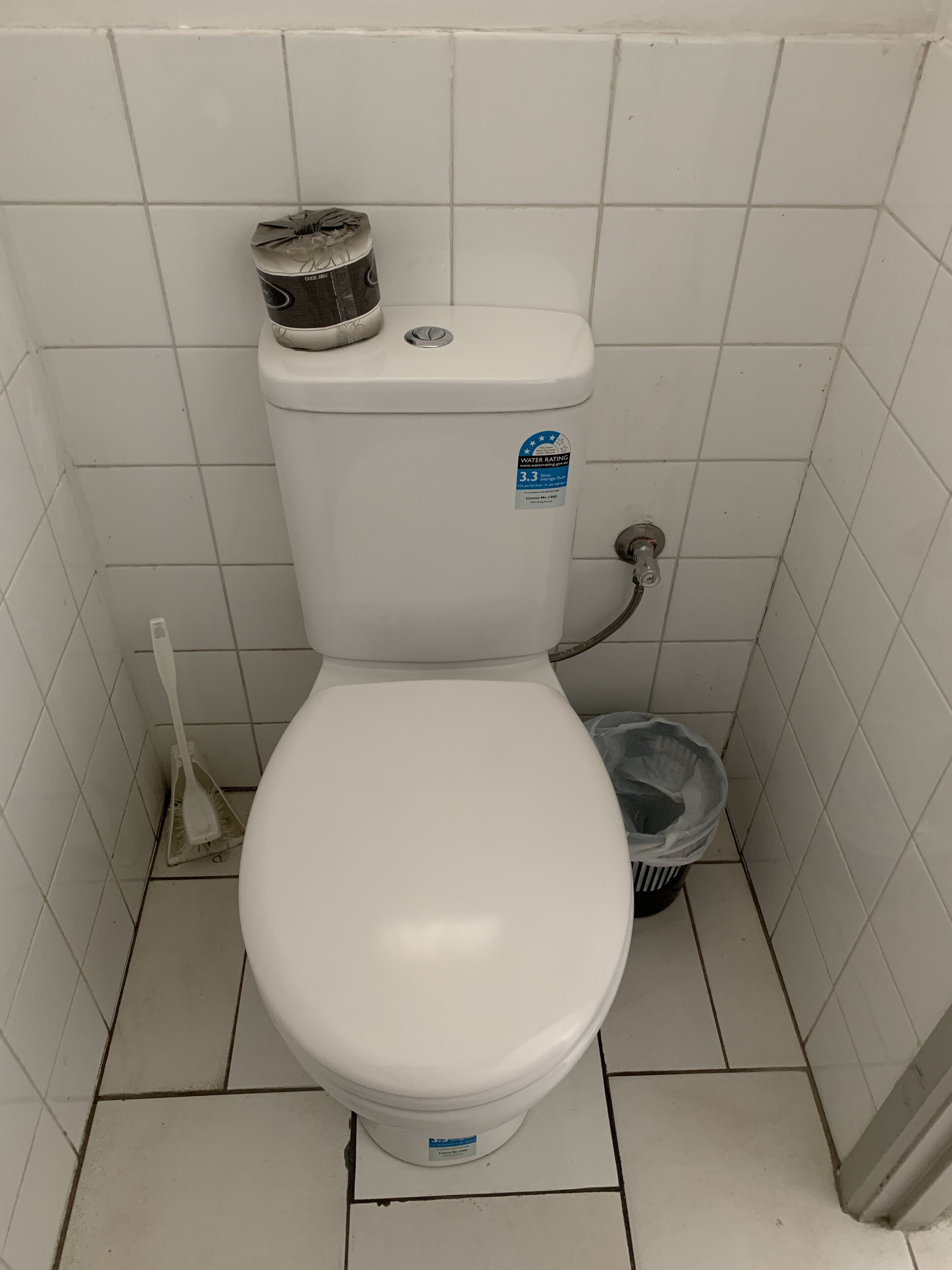 A white toilet with a water rating sticker, a broom, and a trash can in a tiled bathroom.