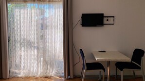 A room with a large window covered by sheer curtains, a flat-screen TV mounted on the wall, a small table, and two chairs. - Beaconsfield Lodge Motel (Beaconsfield)