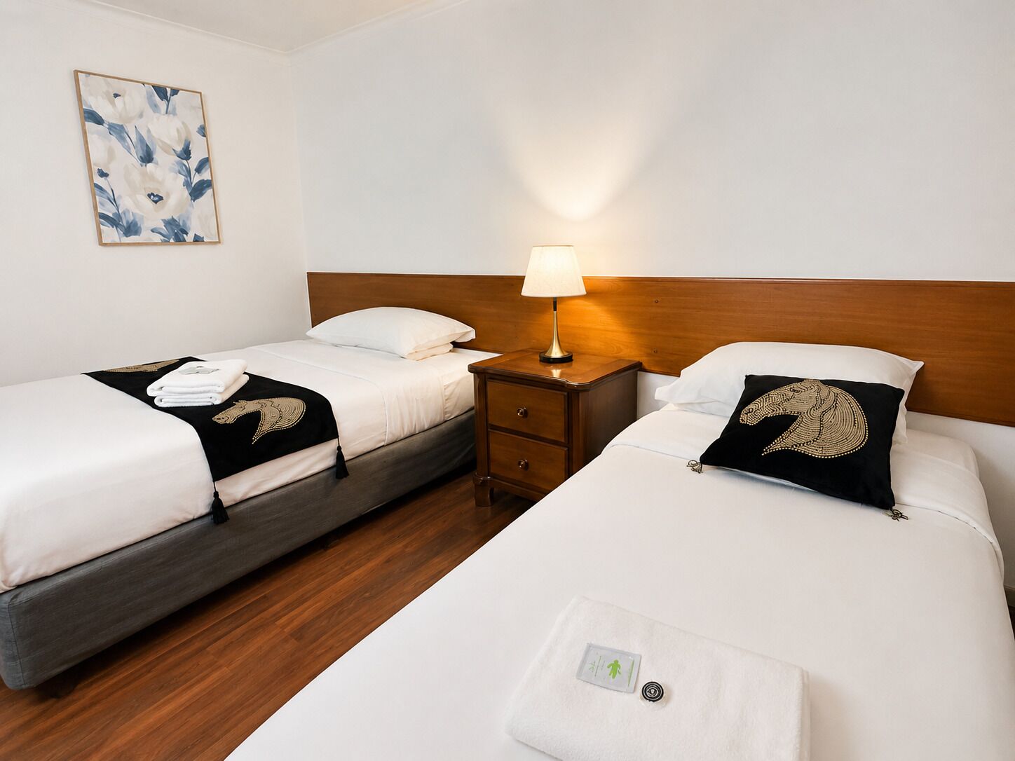A hotel room with two beds, a nightstand with a lamp, a wall-mounted artwork, and a visible towel dispenser.