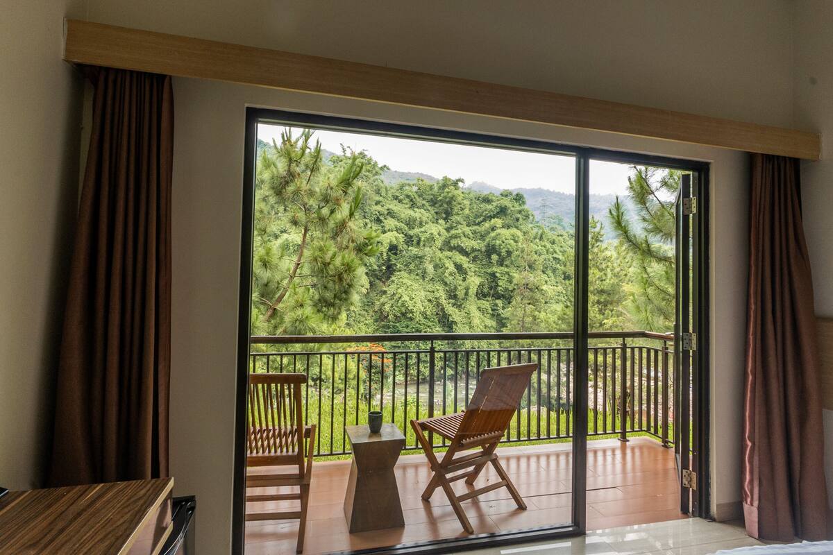A balcony with a view of a lush forest and mountains. There are two wooden chairs and a small table.
