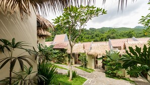 A courtyard with thatched-roof buildings, surrounded by lush greenery and a stone pathway. - Driam Riverside Resort (Pasirjambu)