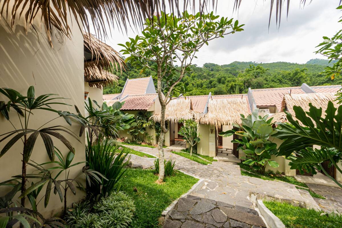 A courtyard with thatched-roof buildings, surrounded by lush greenery and a stone pathway.