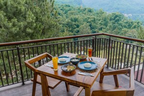 Uma mesa de madeira com uma refeição posta numa varanda com vista para uma área arborizada.