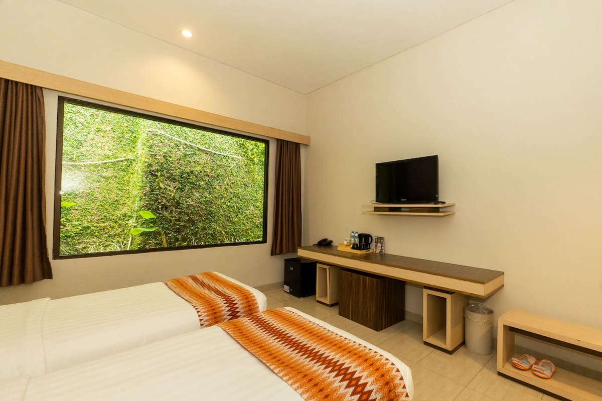 A hotel room with a large window, a bed, a desk with a television, and a small shelf with slippers.