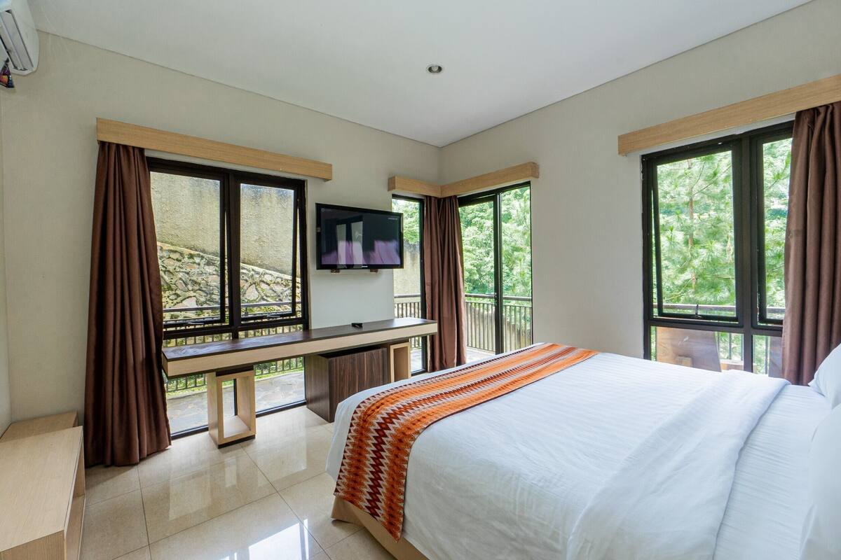 A hotel room with a large bed, a flat-screen TV mounted on the wall, a desk with a chair, and large windows offering a view of greenery.