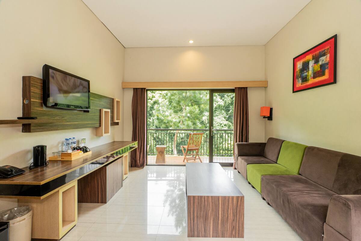 A modern living room with a flat-screen TV mounted on the wall, a sofa, a coffee table, and a balcony with a view of trees.