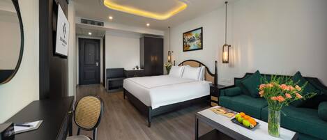 A hotel room with a bed, a green sofa, a desk with a chair, a coffee table with fruit and flowers, and a painting on the wall.