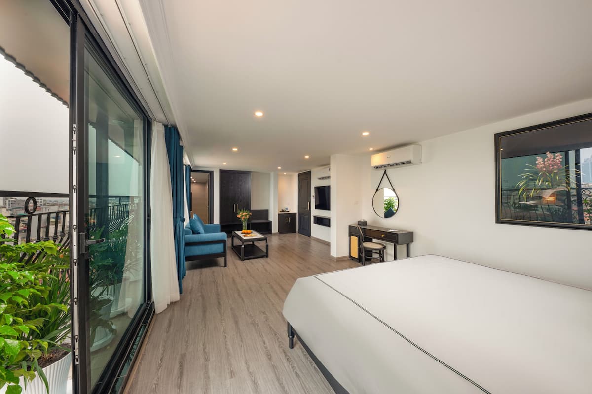 A modern hotel room with a large bed, a seating area with a blue sofa and coffee table, a small desk, and a balcony with plants.