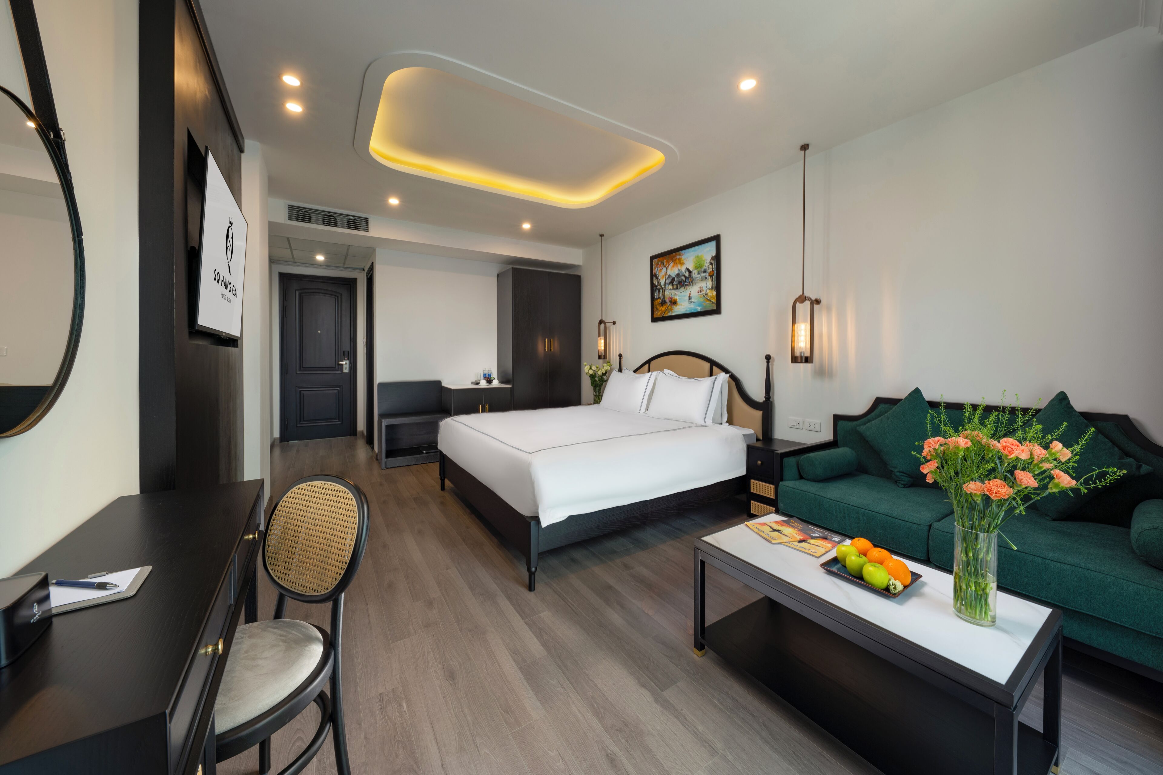 A hotel room with a bed, a green sofa, a desk with a chair, a coffee table with fruit and flowers, and a painting on the wall.