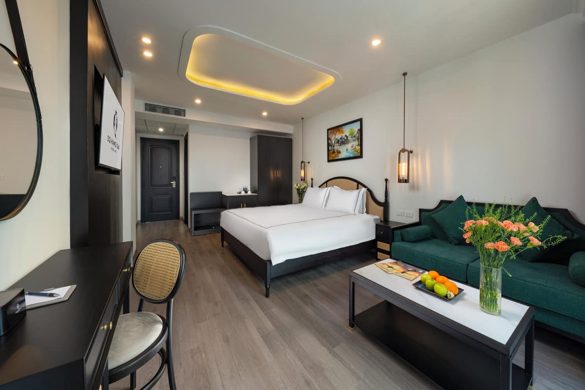 A hotel room with a bed, a green sofa, a desk with a chair, a coffee table with fruit and flowers, and a painting on the wall.