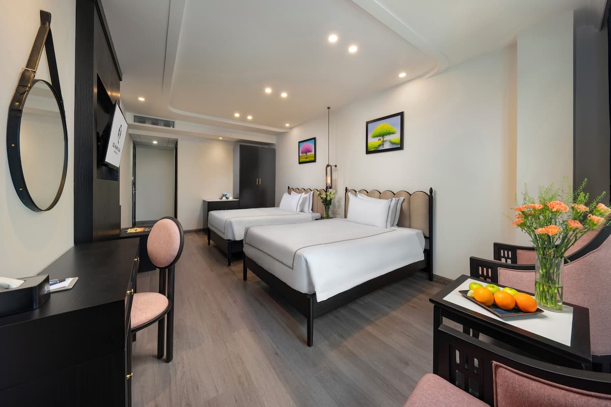 A hotel room with two beds, a desk, a chair, a small table with fruit and flowers, and artwork on the walls.