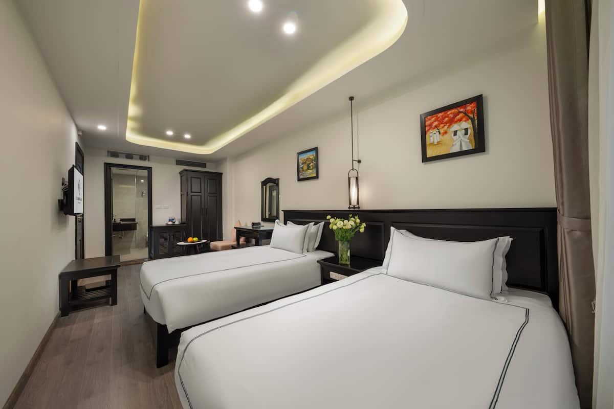 A hotel room with two beds, a bedside table, a nightstand, a mirror, a painting, and a ceiling light fixture.