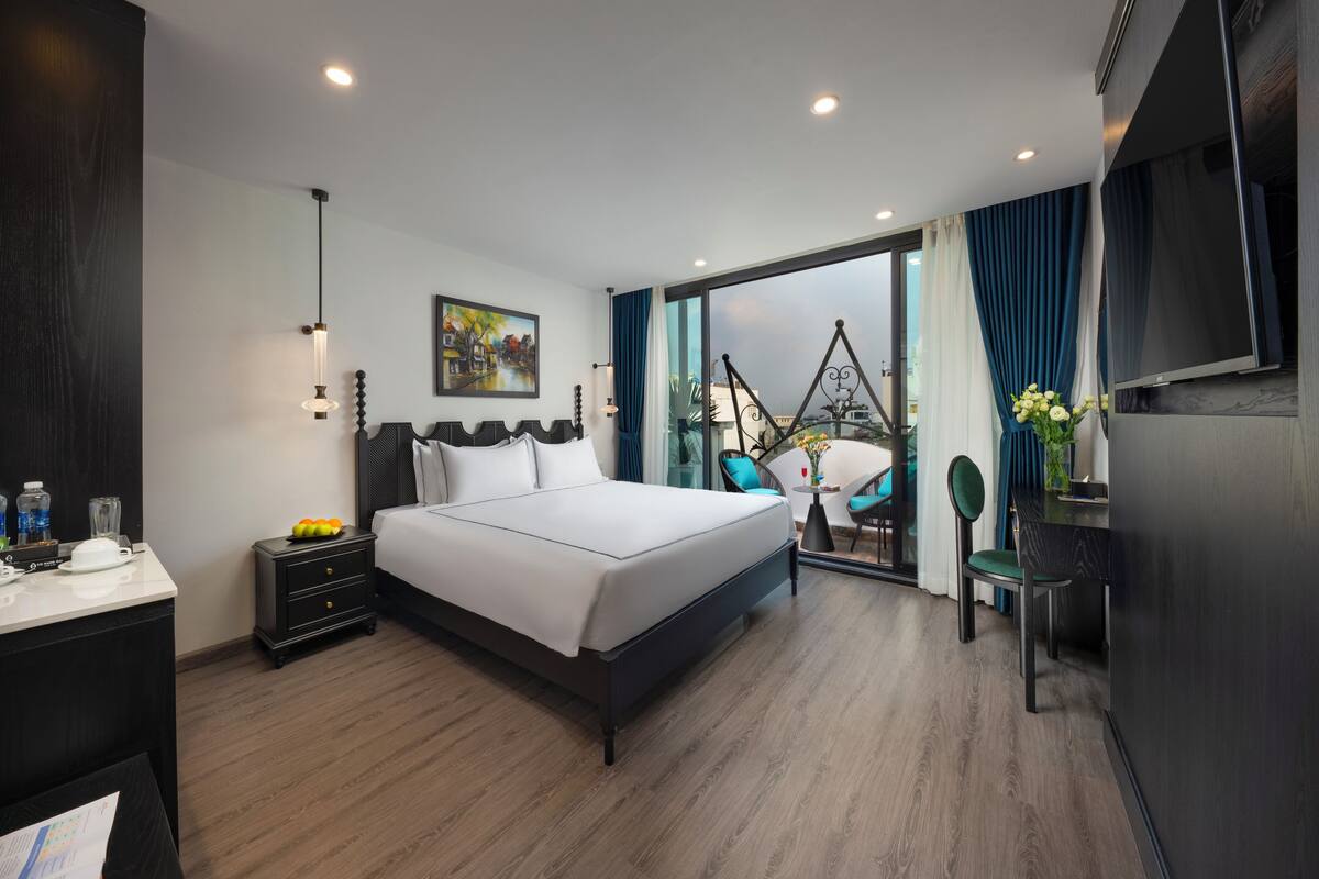 A modern hotel room with a large bed, a desk with a chair, a TV, and a balcony with a view.