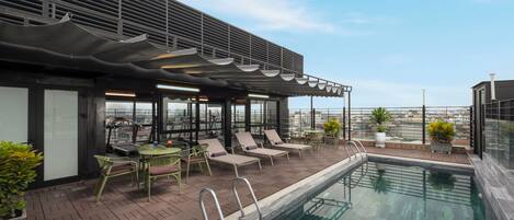 A rooftop pool area with lounge chairs and a table, surrounded by a modern building with large windows.