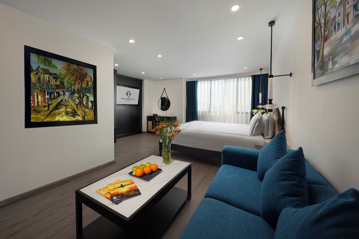 A modern hotel room with a bed, a blue sofa, a coffee table with fruits and a magazine, a painting on the wall, and a view of the street through the window.