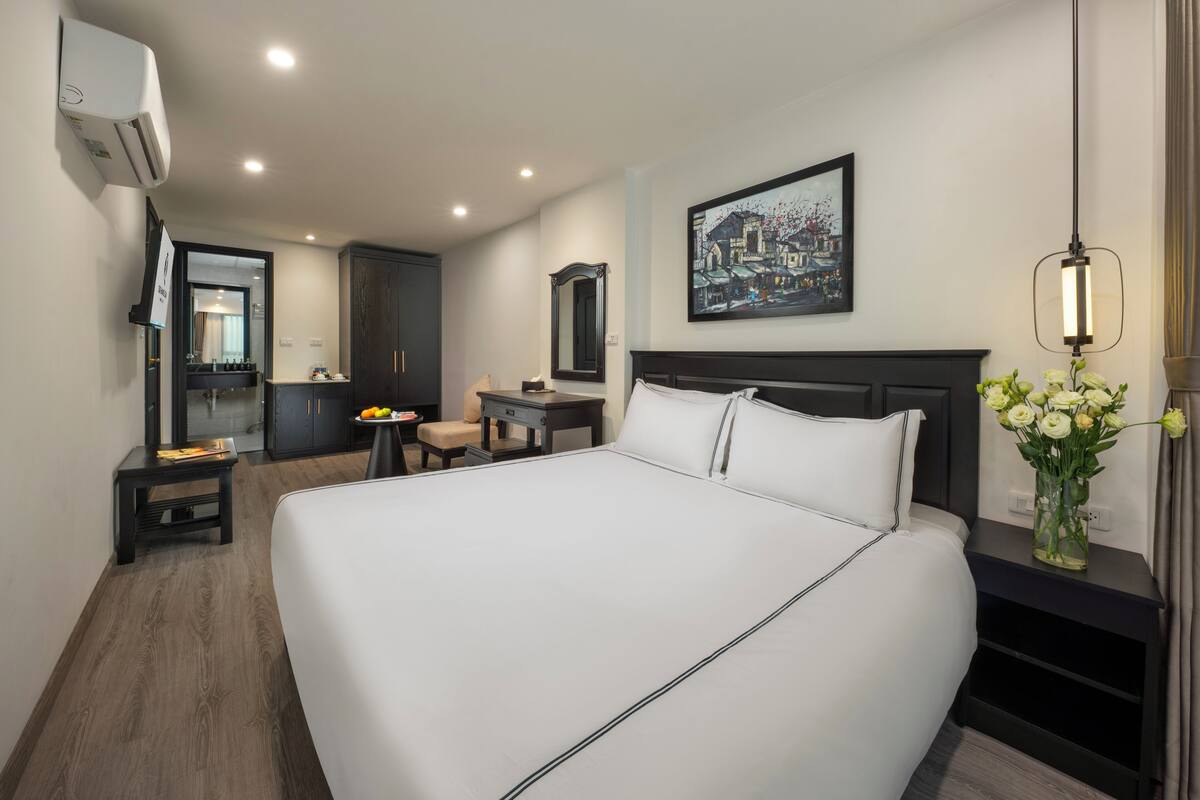 A hotel room with a large bed, a desk, a chair, a vanity, and a painting on the wall.