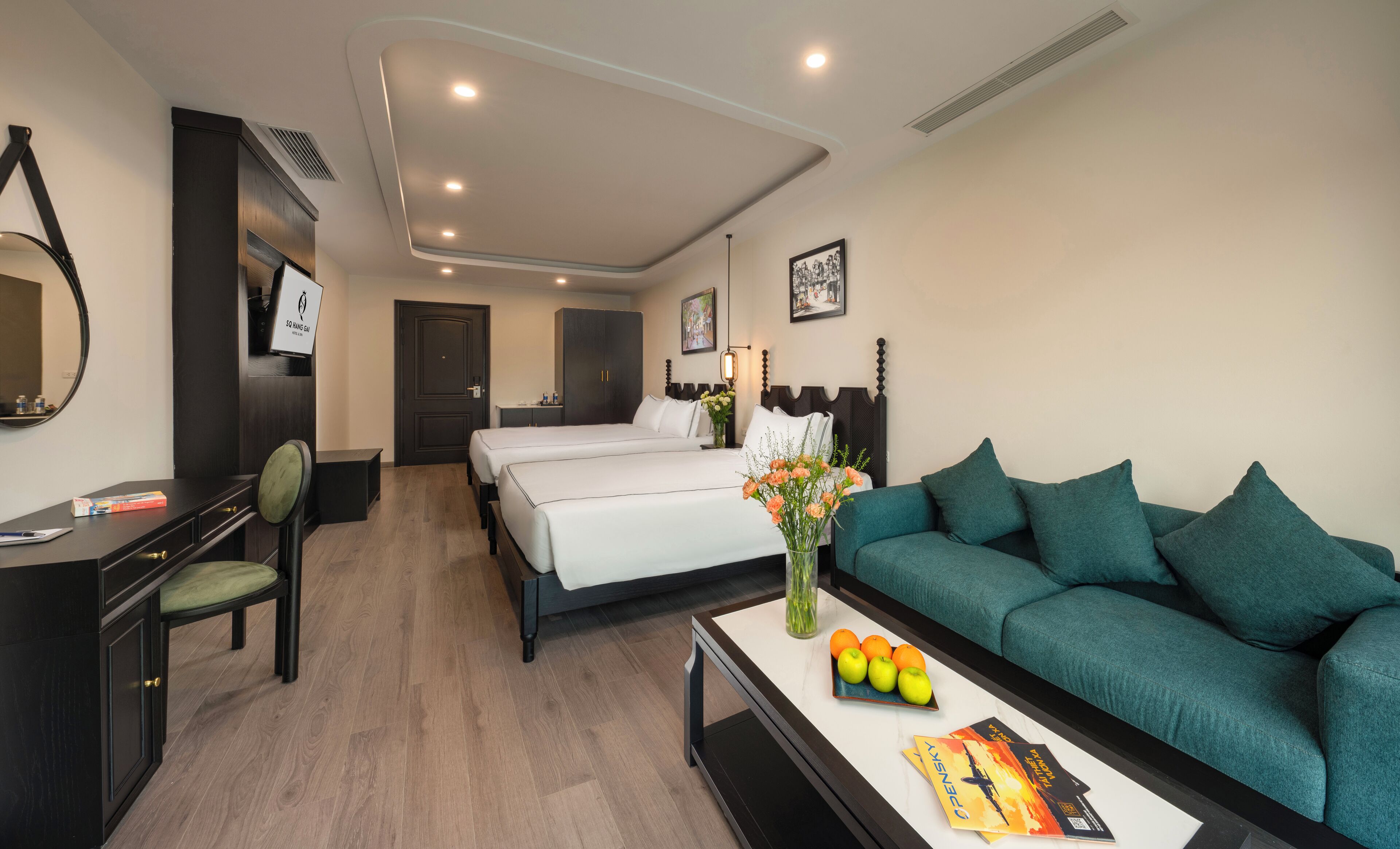 Habitación de hotel con cama, escritorio, silla, sofá y mesa de centro con libros y fruta.