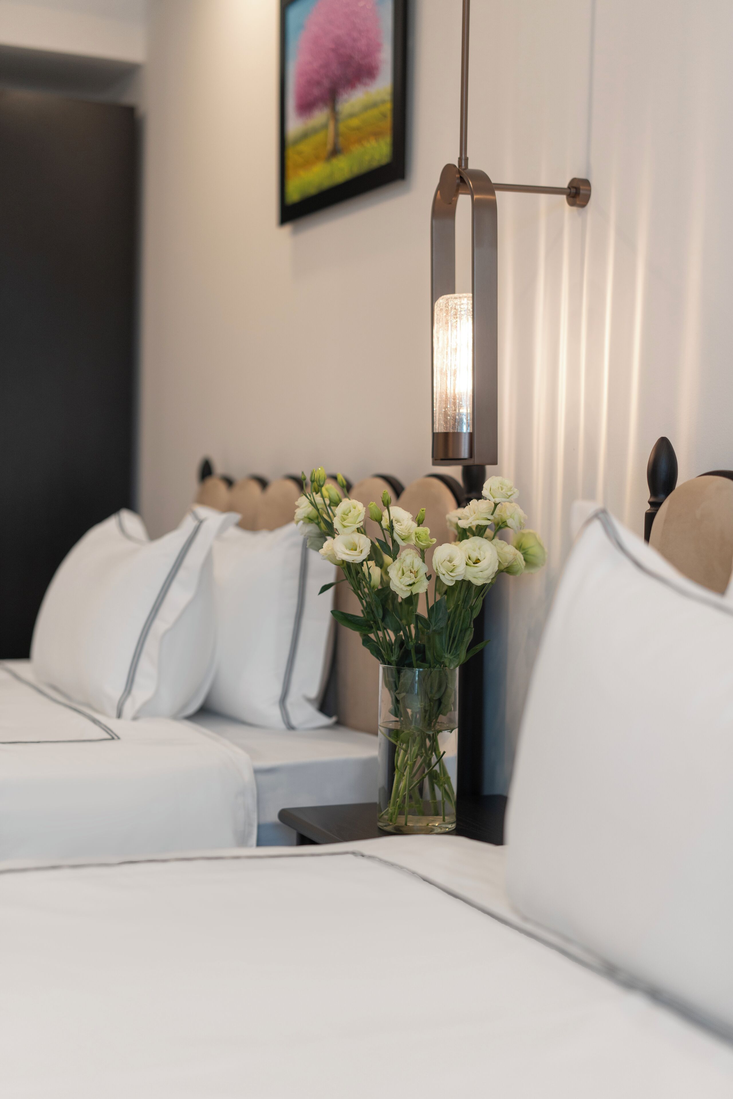 A hotel room with a bed, a bedside table with a vase of flowers, and a wall-mounted light fixture.
