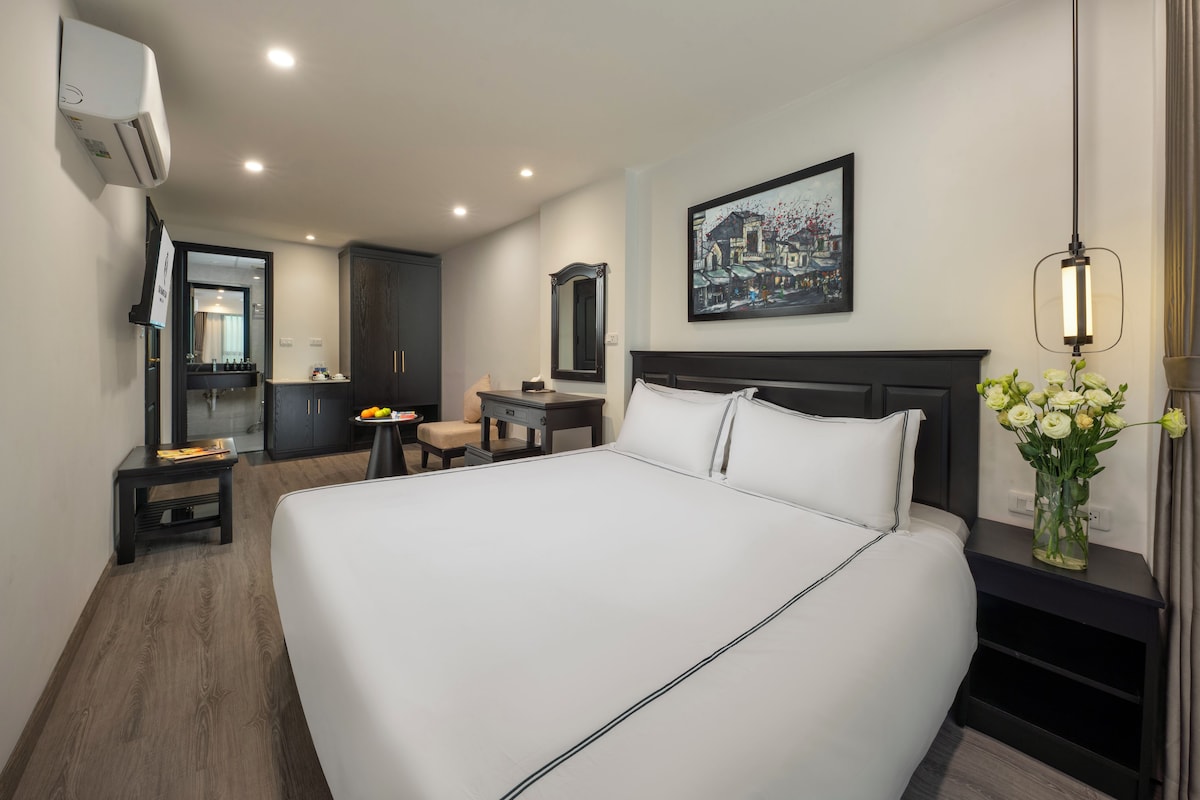 A hotel room with a large bed, a desk, a chair, a vanity, and a painting on the wall.
