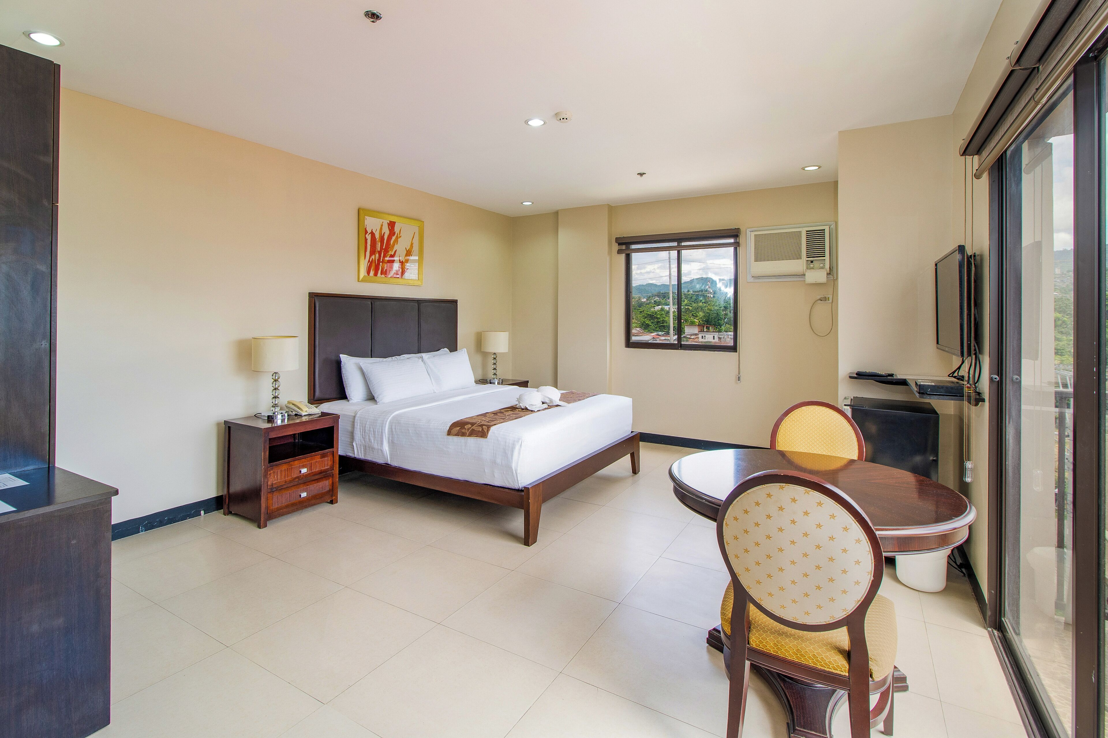 A hotel room with a bed, bedside table, lamp, chair, dining table, and a TV.
