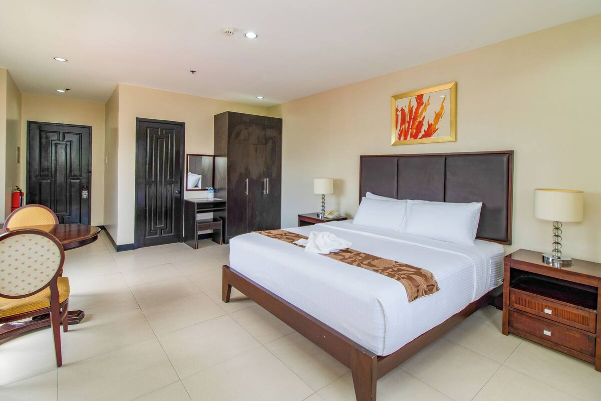 A hotel room with a large bed, a desk with a chair, a wardrobe, and a bedside table with a lamp.