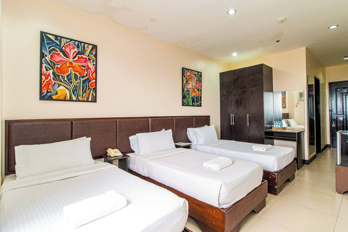 A hotel room with three beds, a desk, a wardrobe, and two paintings on the wall.