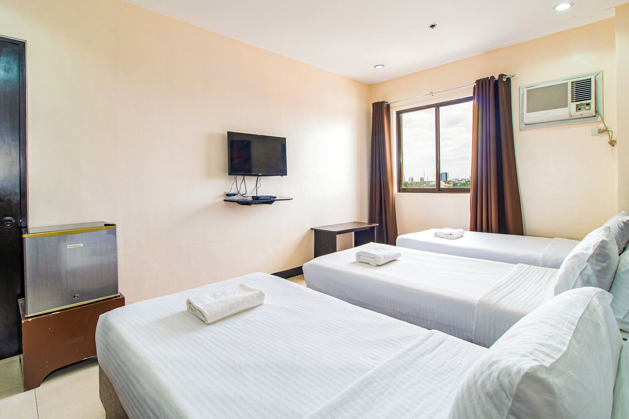 A hotel room with two beds, a TV, an air conditioning unit, and a window with curtains.
