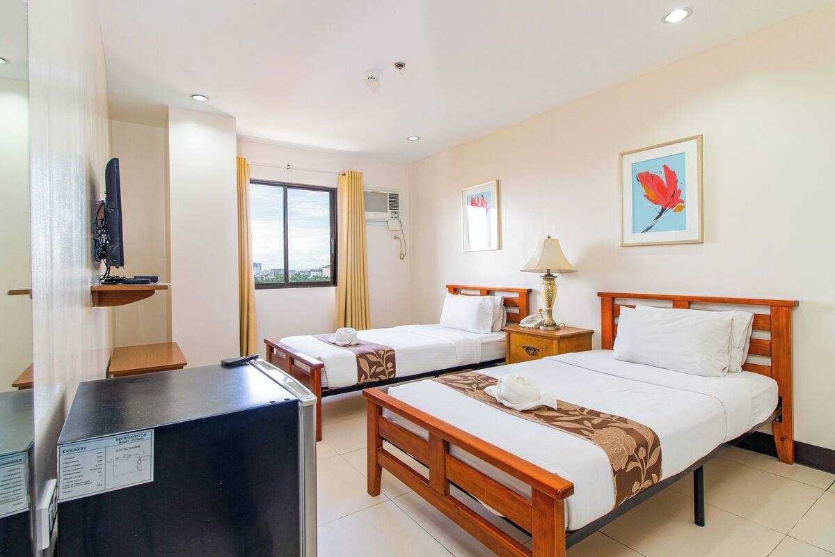 A hotel room with two beds, a desk, a TV, and a window with curtains.