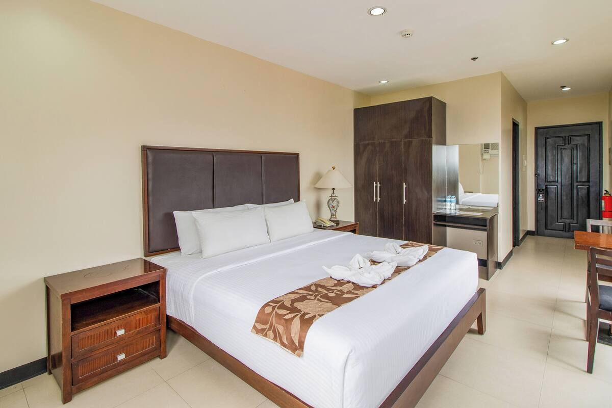 A hotel room with a large bed, a wooden nightstand, a desk with a chair, a wardrobe, and a bathroom visible through an open door.