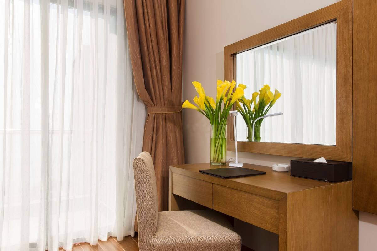 A hotel room with a wooden desk, a chair, a vase with yellow flowers, and a large mirror.