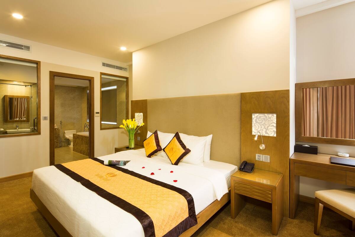 A hotel room with a large bed, a desk, a chair, a bedside table, a mirror, and a bathroom visible through an open door.