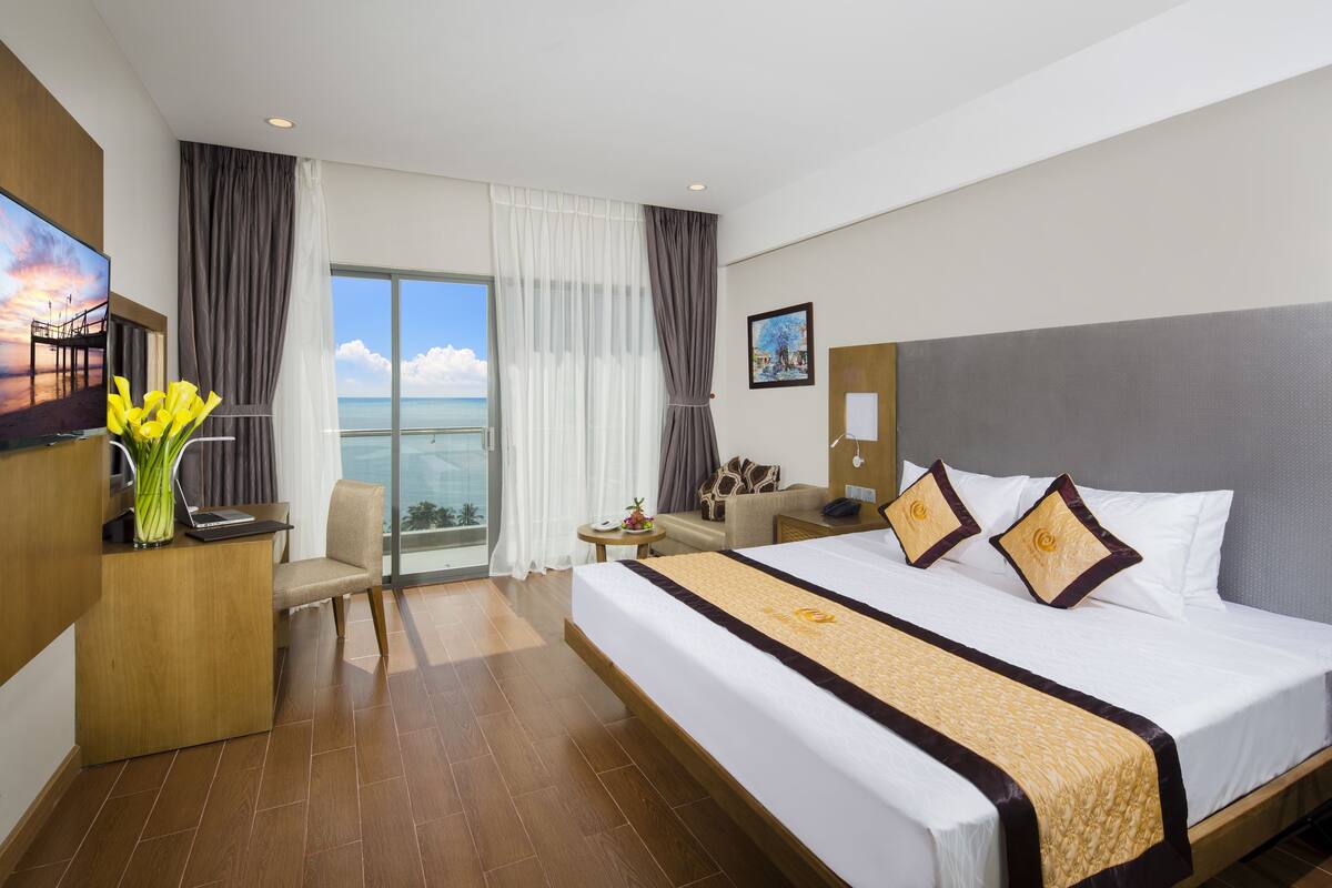 A hotel room with a large bed, a desk with a laptop, a chair, a sofa, a TV, and a view of the ocean.